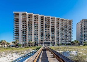 Un hotel de gran altura con múltiples balcones, un paseo marítimo de madera que conduce a la entrada y un cielo azul despejado.