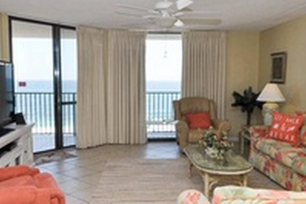 Habitación con sofá, sillón y televisor sobre un mueble bajo. Se ve una ventana con cortinas, un ventilador de techo y vista al mar.