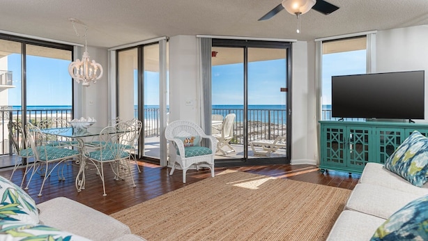 A spacious living room with a large TV, a dining area with white chairs, and a balcony with a view of the ocean. - Phoenix East by Brett Robinson Vacations (Orange Beach)