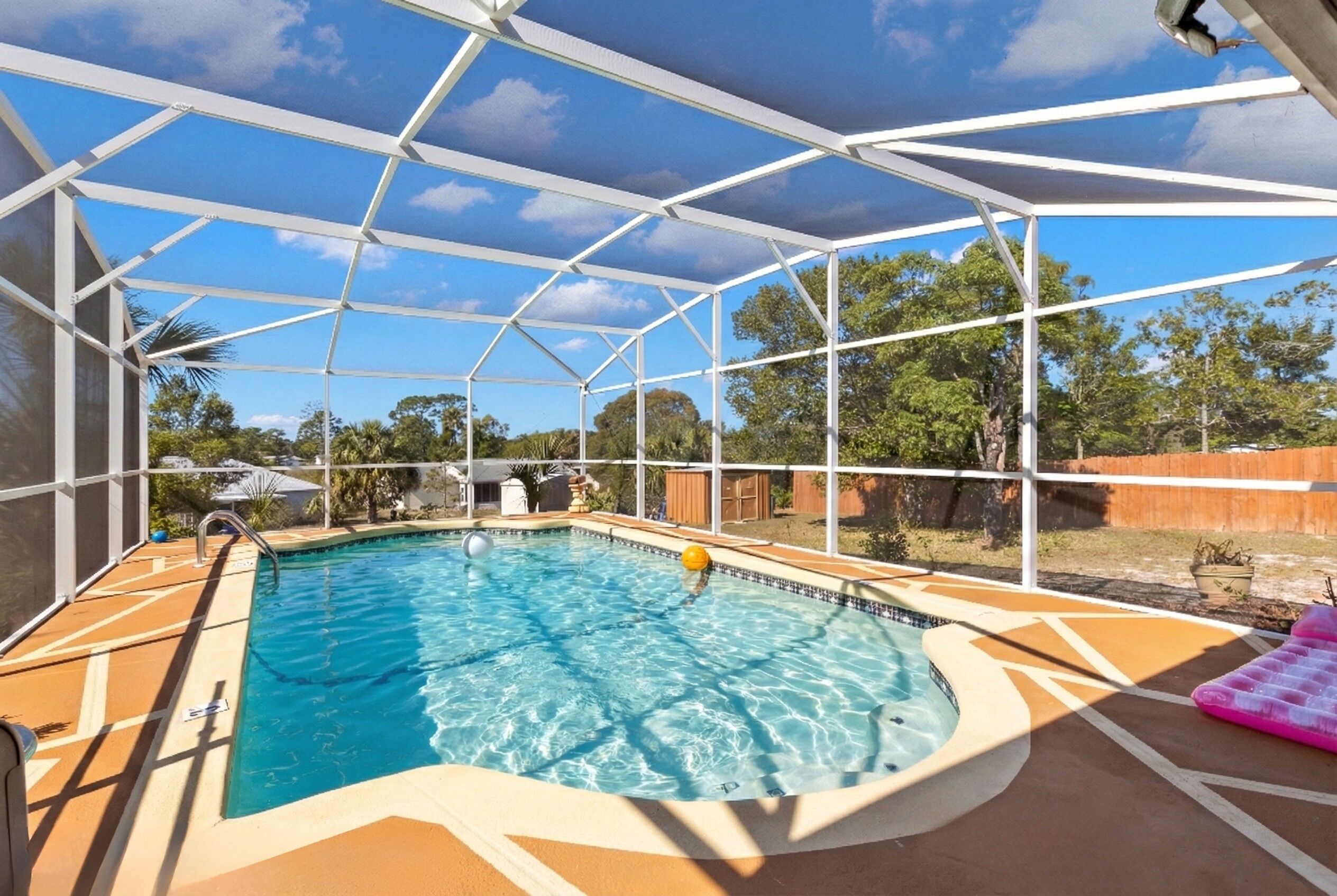 Una piscina techada con agua azul cristalina, rodeada por una valla blanca y un sillón inflable rosa a la derecha.