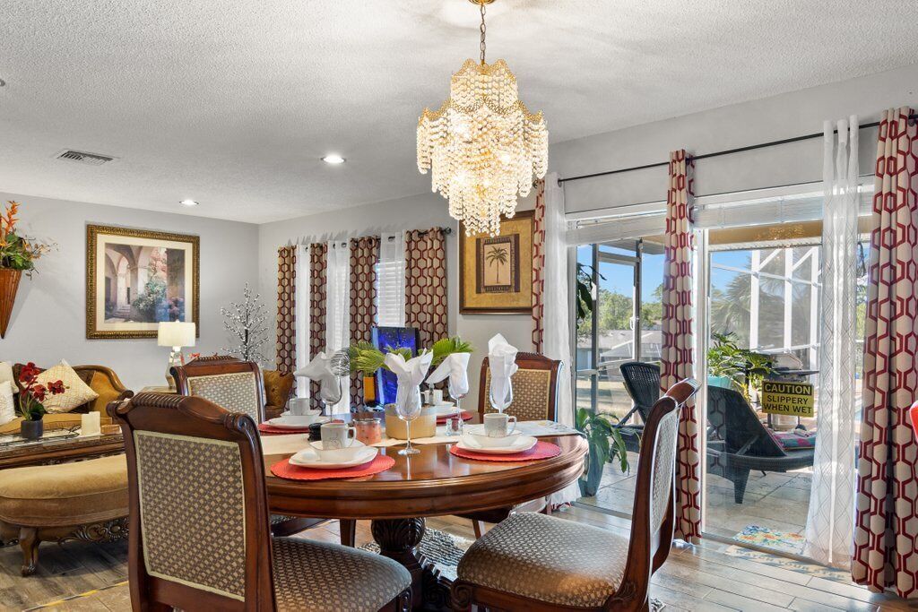 A dining area with a round table set for four, surrounded by upholstered chairs with a patterned fabric. A chandelier hangs from the ceiling, and the room has a large window with patterned curtains.