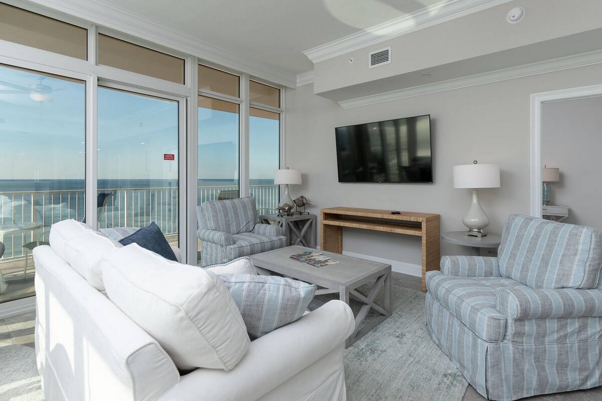 A modern living room with a balcony overlooking the ocean, featuring a white sofa, striped armchairs, a coffee table, and a TV.