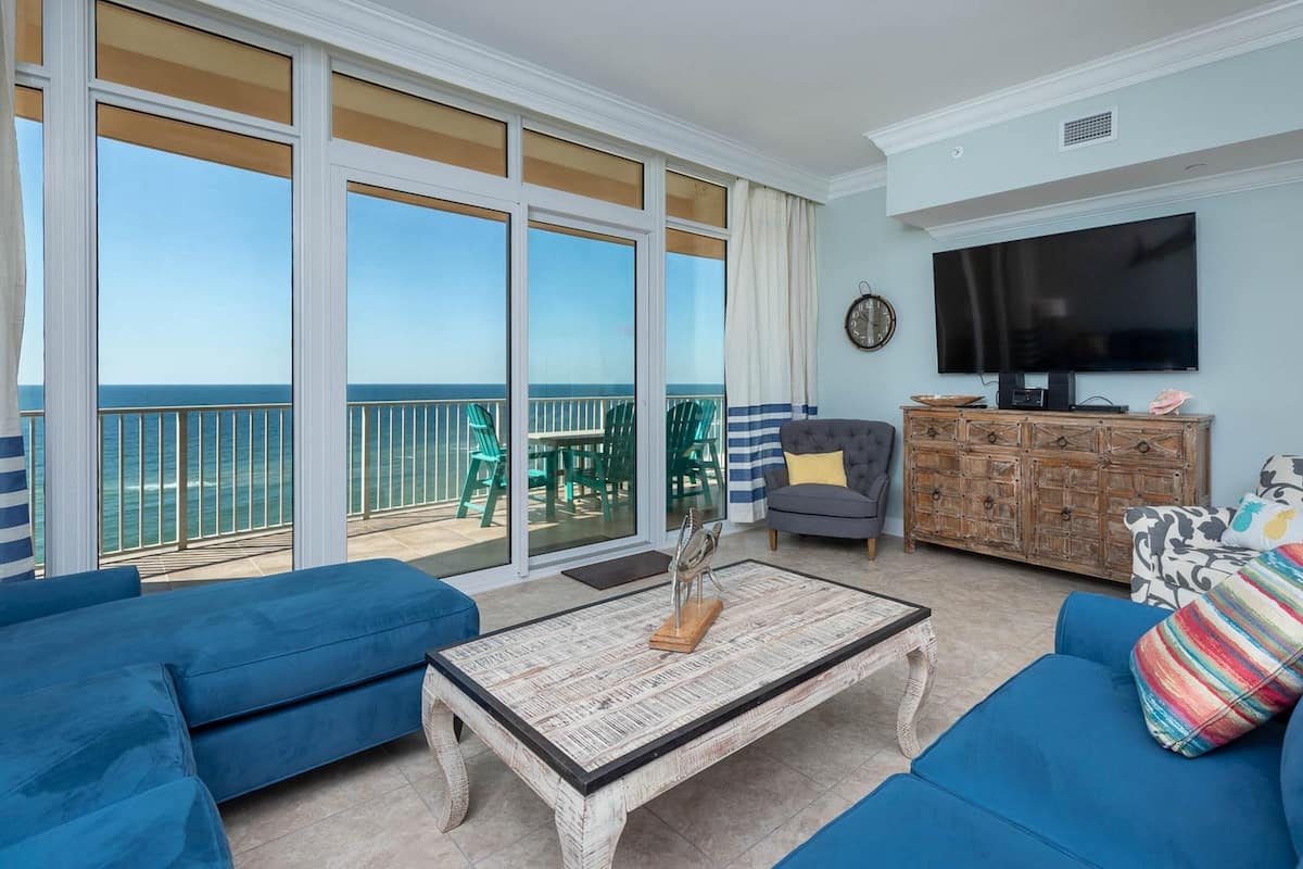 A spacious living area with a large sliding glass door leading to a balcony with a view of the ocean. There is a television mounted on the wall, a wooden cabinet, and two blue sofas.