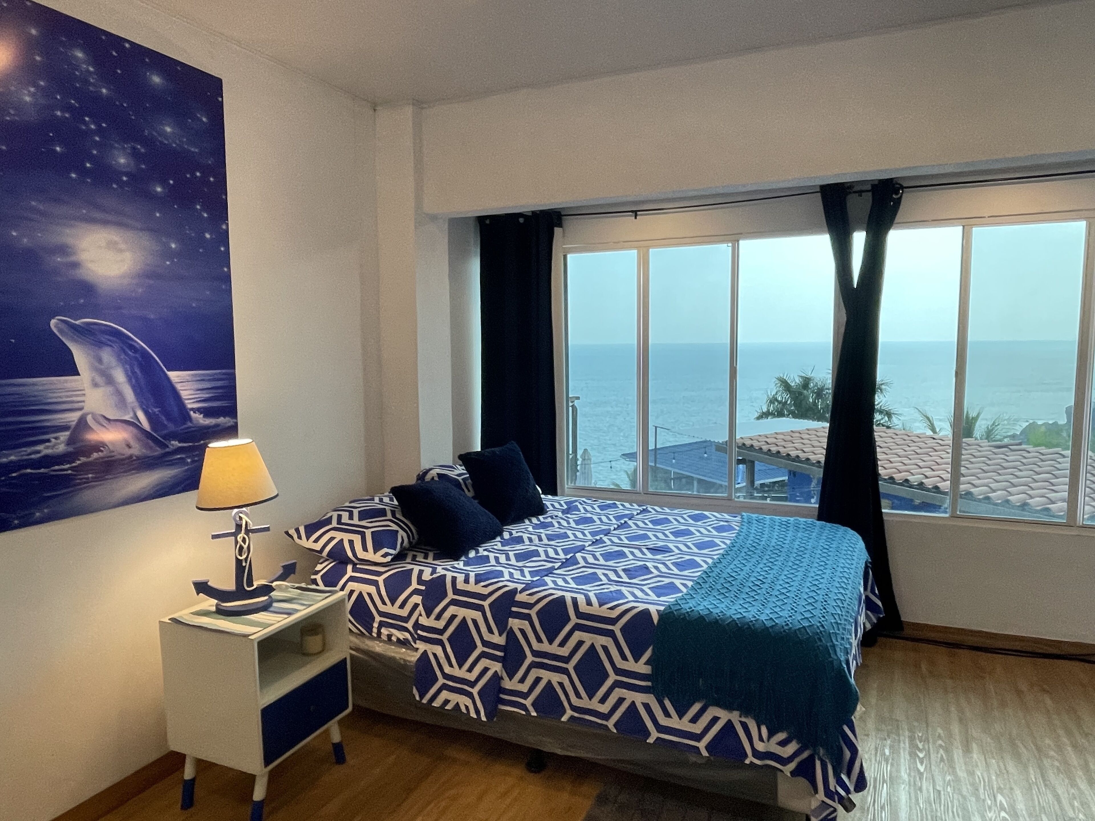 Un dormitori amb llit, una tauleta de nit, una làmpada, un quadre d'una balena i vista al mar per la finestra.
