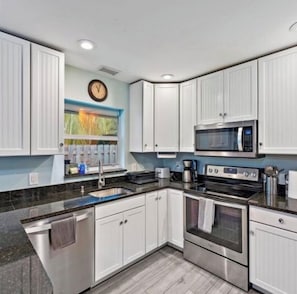 A modern kitchen with white cabinets, a stainless steel oven, a microwave, and a window with a view of greenery. - Singer Island pool home (West Palm Beach)