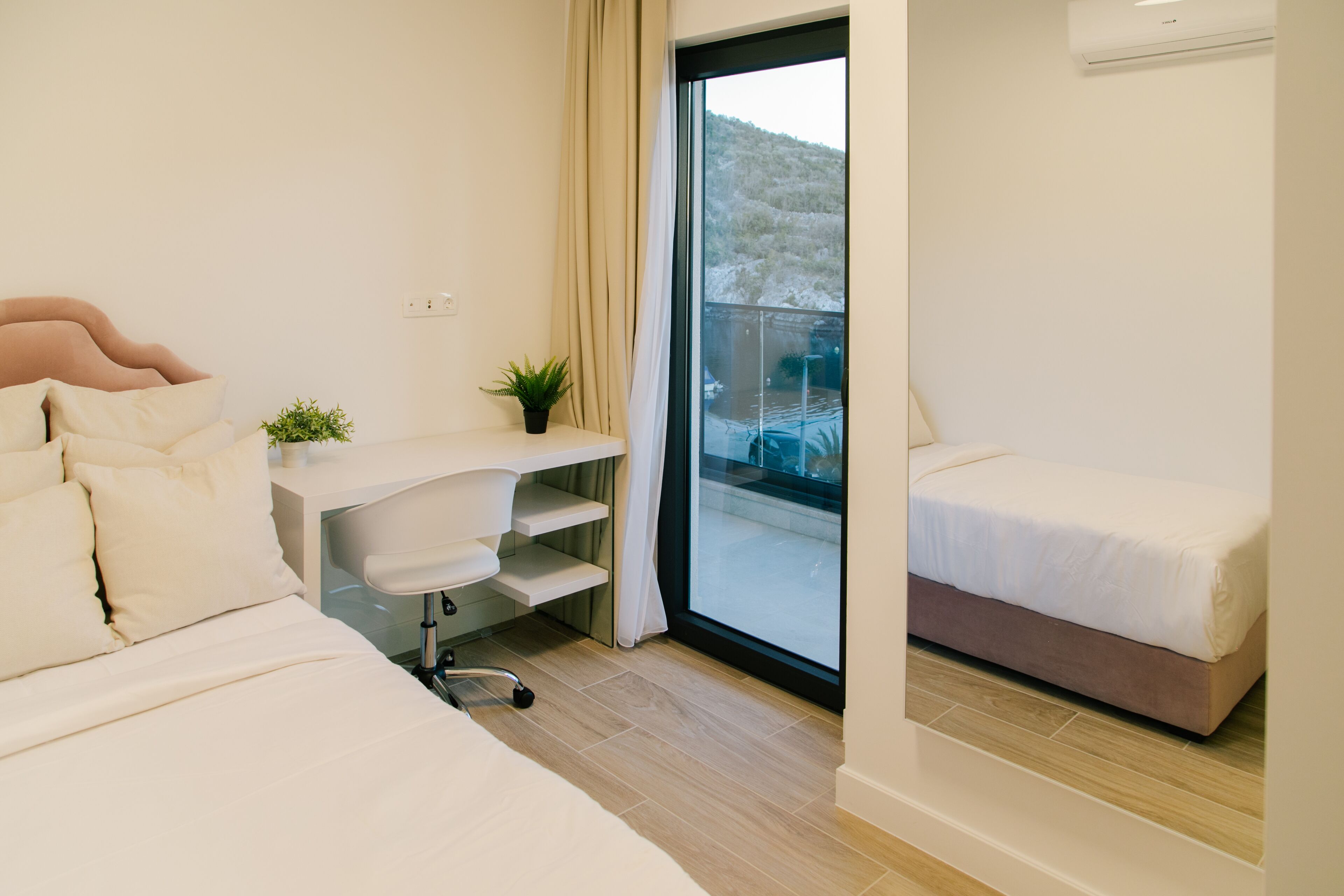Ein modernes Hotelzimmer mit einem großen Bett, einem Schreibtisch mit Stuhl, einem kleinen Balkon und einer Zimmerpflanze.