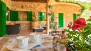 Ein gemütlicher Sitzbereich im Freien mit Steinmauern und grünen Fensterläden, mit einem Tisch, Tassen und einem Topf mit roten Blüten.