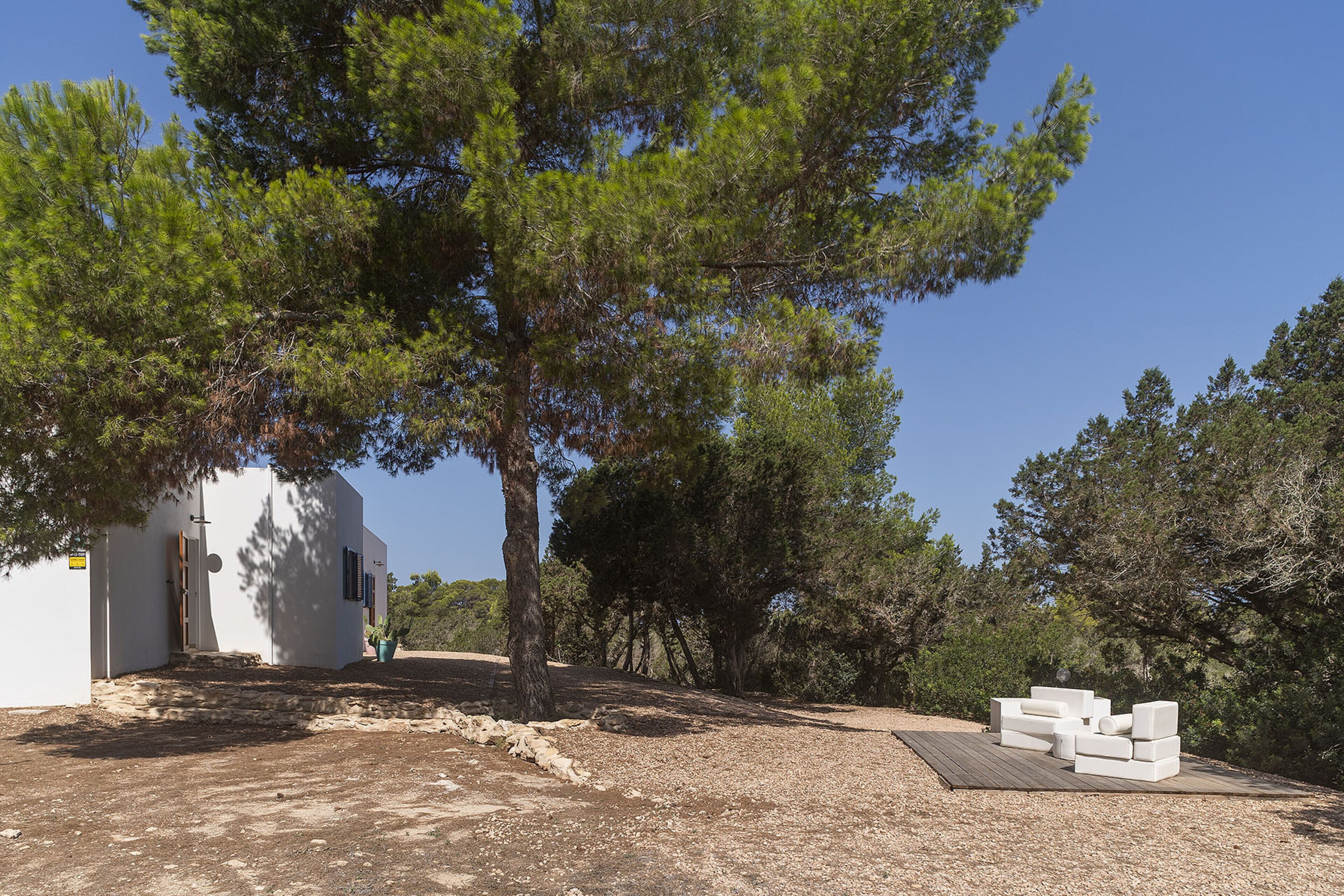 Un edificio moderno con fachada blanca, rodeado de árboles y un área de grava con mobiliario exterior blanco.
