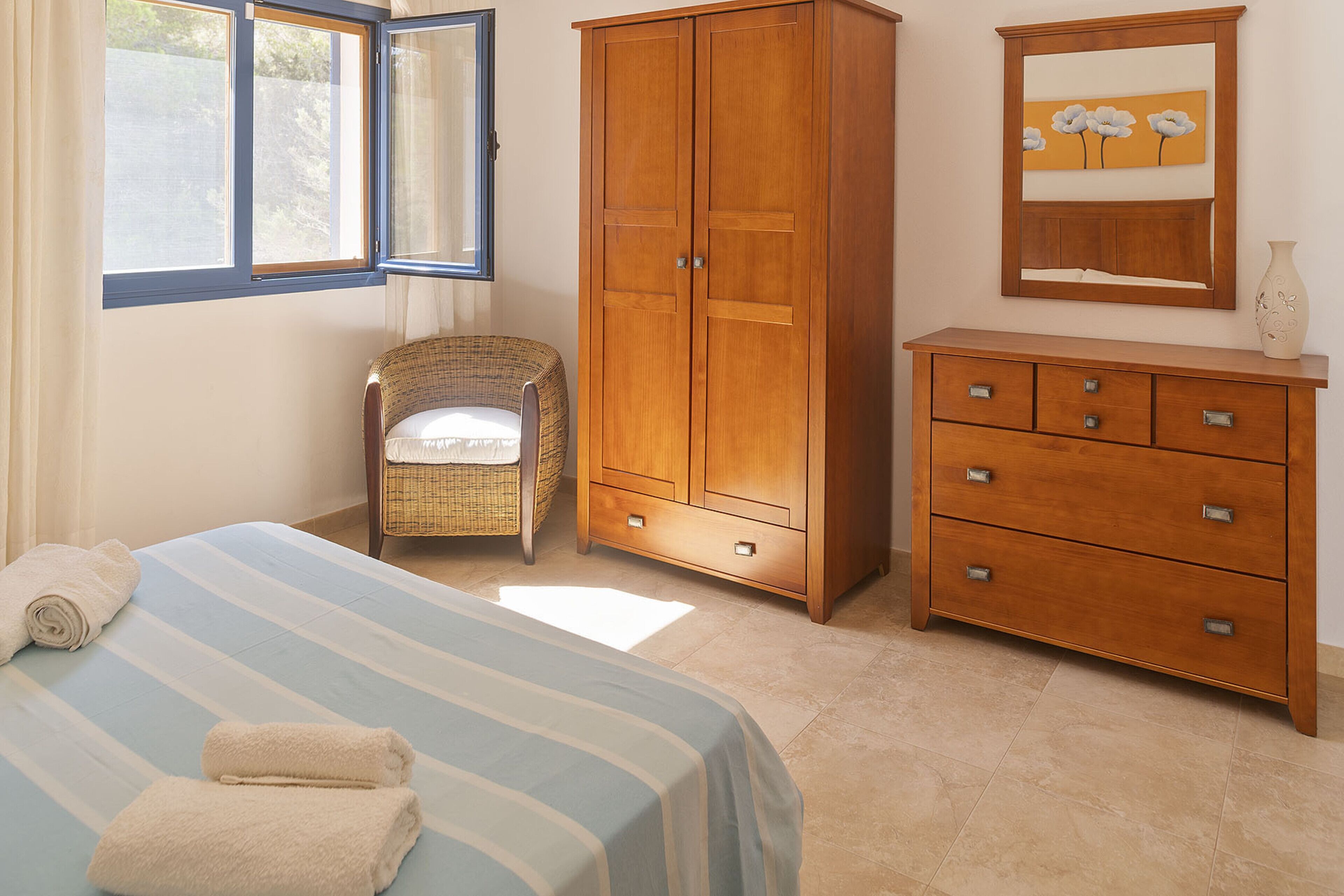 A bedroom with a bed, a wooden wardrobe, a wicker chair, and a dresser.