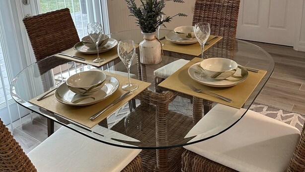 Comedor con una mesa de vidrio rodeada de sillas de mimbre con cojines. Hay una planta en maceta sobre la mesa y un reloj de pared encima.