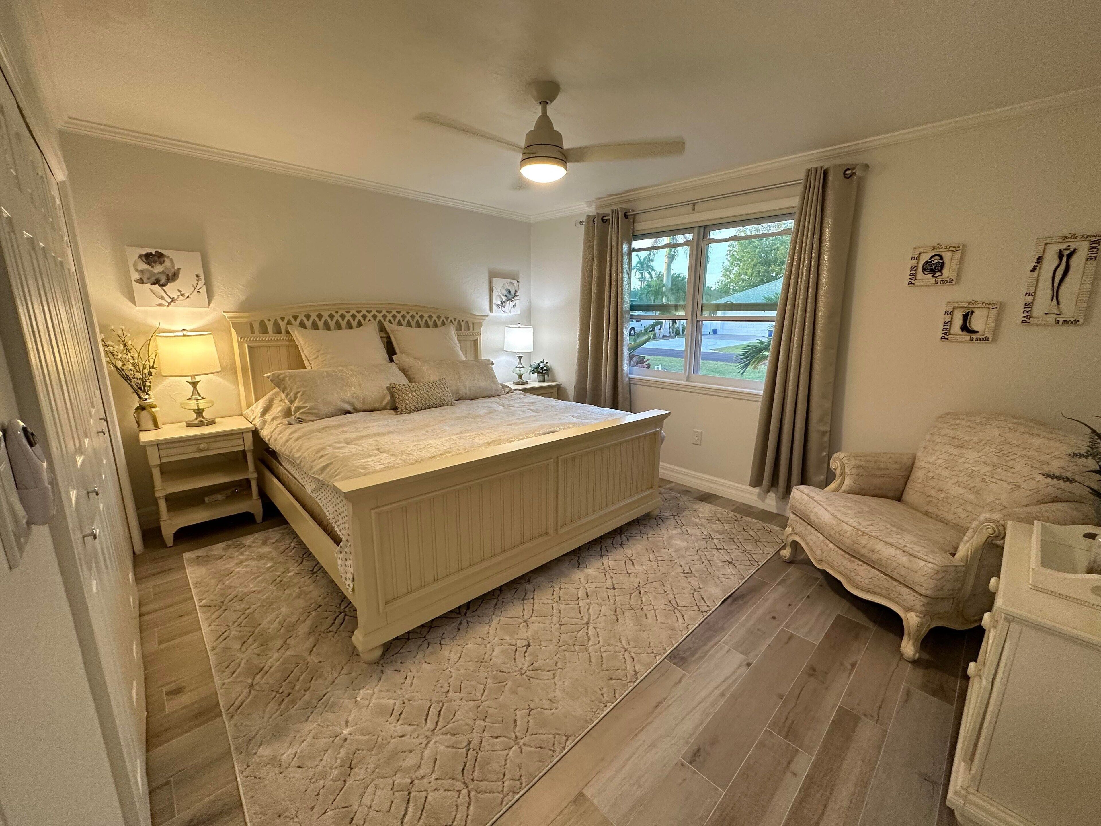 Ein Schlafzimmer mit einem großen Bett, einem Nachttisch, einem Sessel, einem Deckenventilator und einem Fenster mit Vorhängen.