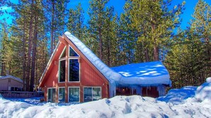 A red cabin with a steeply pitched roof, surrounded by snow and tall pine trees.