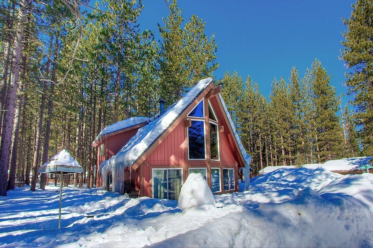 A red cabin with large windows and a snow-covered roof, surrounded by tall pine trees.