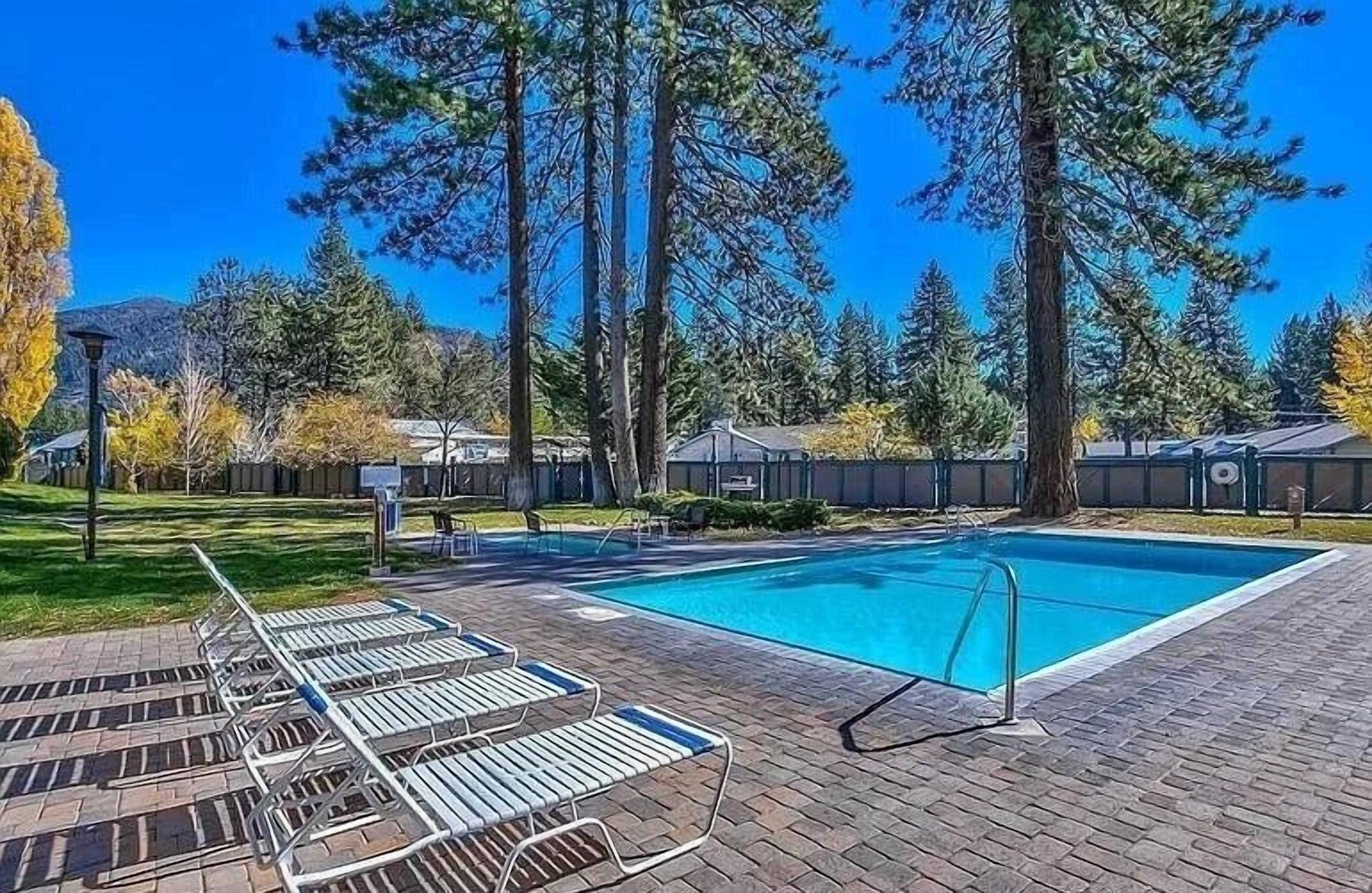 A rectangular swimming pool with a clear blue water, surrounded by a paved area with lounge chairs and a brick pathway.