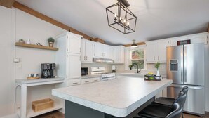 Une cuisine avec des meubles blancs, un grand îlot central, un réfrigérateur en acier inoxydable et un luminaire suspendu.