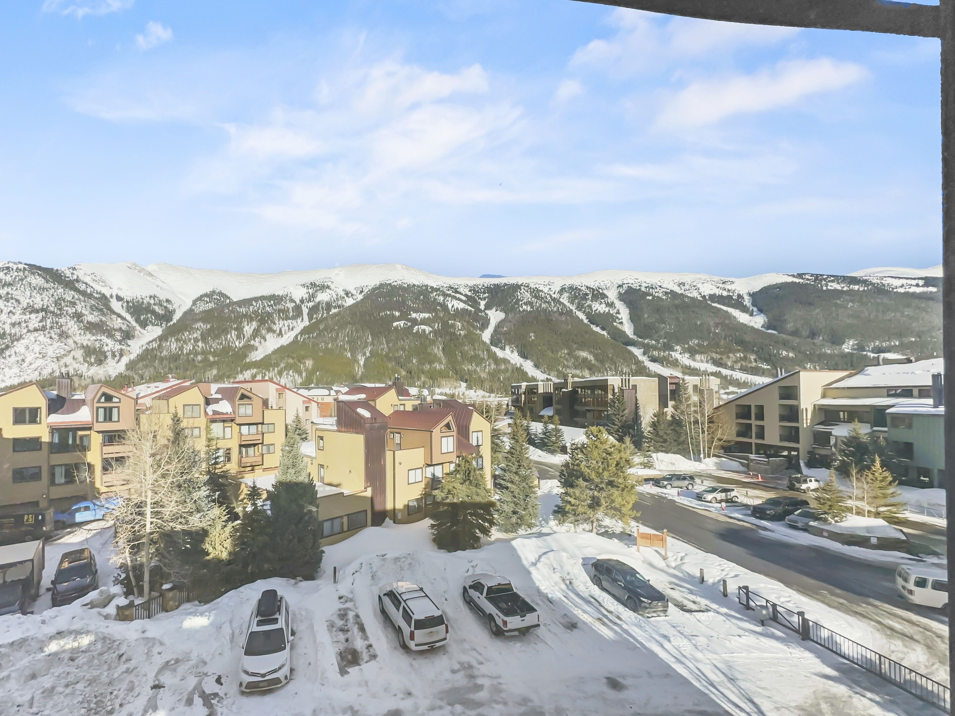 A view of a residential area with snow-covered ground, parked cars, and buildings with a modern architectural style.
