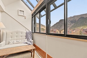 A room with a sofa, a wooden table, and a view of houses and mountains through large windows.