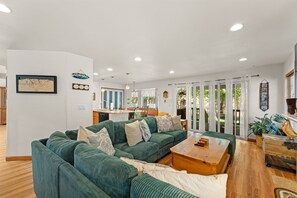 A spacious living room with a green sectional sofa, a wooden coffee table, and a kitchen area visible through a glass door. - Wolfpack Refuge 4 Bedroom Home by RedAwning (South Lake Tahoe)