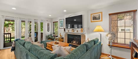 A modern living room with a sectional sofa, a fireplace, a television, and a large window with blinds.