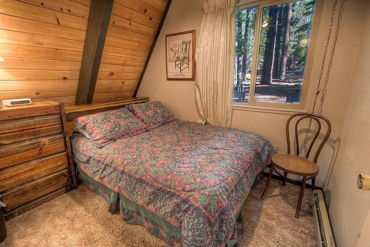 Une chambre avec un plafond en pente en bois, un lit recouvert d’une couvre-lit à motifs, une chaise, une commode et une fenêtre donnant sur les arbres.
