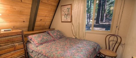 Ein Schlafzimmer mit holzgetäfelter Decke, einem Bett mit gemustertem Überwurf, einem Sessel, einer Kommode und einem Fenster mit Blick auf Bäume.