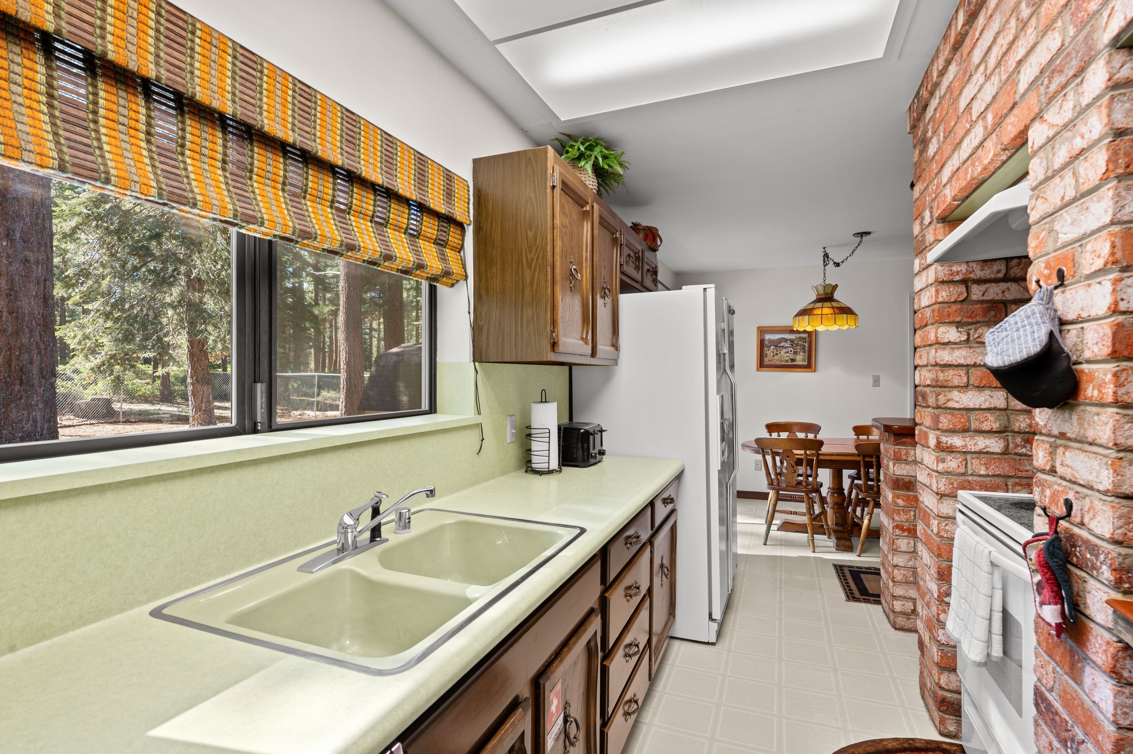 Een keuken met een groot raam, houten kasten, een koelkast en een eetgedeelte met een bakstenen muur en een hangende pannendrager.