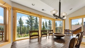 Un comedor con una gran mesa de madera, varias sillas y un tazón. La habitación tiene ventanas amplias con vistas a árboles y montañas.