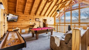 Interior de una cabaña de madera con mesa de billar, un sofá y una televisión instalada en la pared.