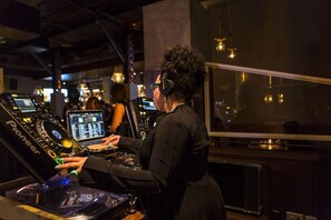 Une personne manipule du matériel de DJ dans une pièce faiblement éclairée, où l’on aperçoit des écrans et une grande fenêtre.