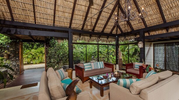 Un espacio interior amplio con techo de paja, asientos cómodos y vistas a exuberante vegetación.