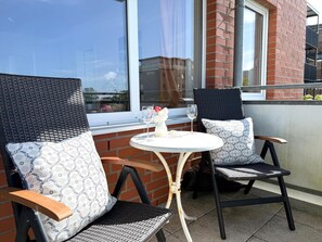 Two wicker chairs with patterned cushions, a small round table, and a glass of wine on a balcony with a brick wall and windows.