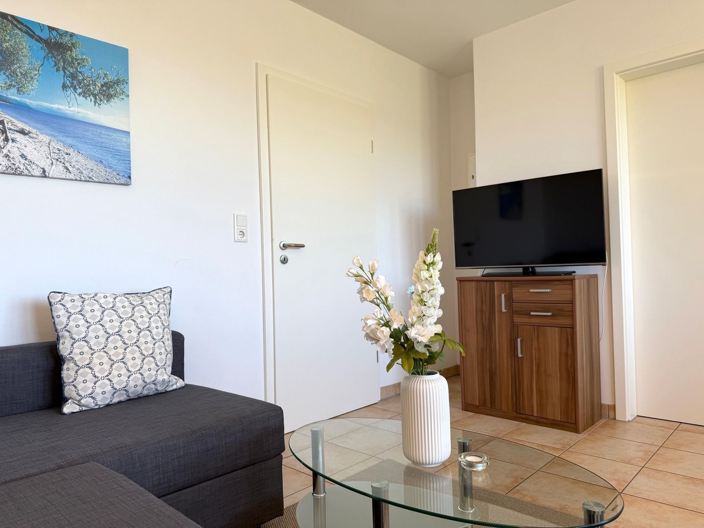Ein Wohnzimmer mit einer Couch, einem Glastisch, einer Blumenvase, einem Fernseher und einer Holzkommode.