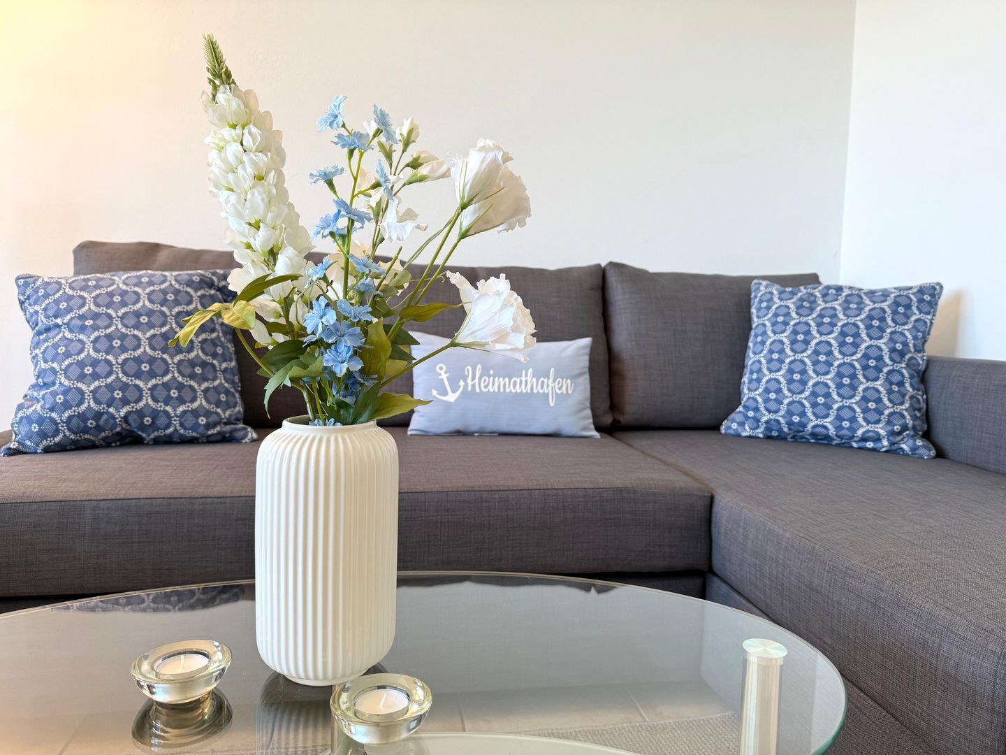 Ein modernes Wohnzimmer mit einem Glastisch, einer weißen Vase mit Blumen und gemusterten Kissen auf einem grauen Sofa.