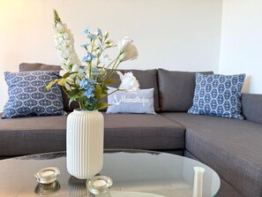 Ein modernes Wohnzimmer mit einem Glastisch, einer weißen Vase mit Blumen und gemusterten Kissen auf einem grauen Sofa.
