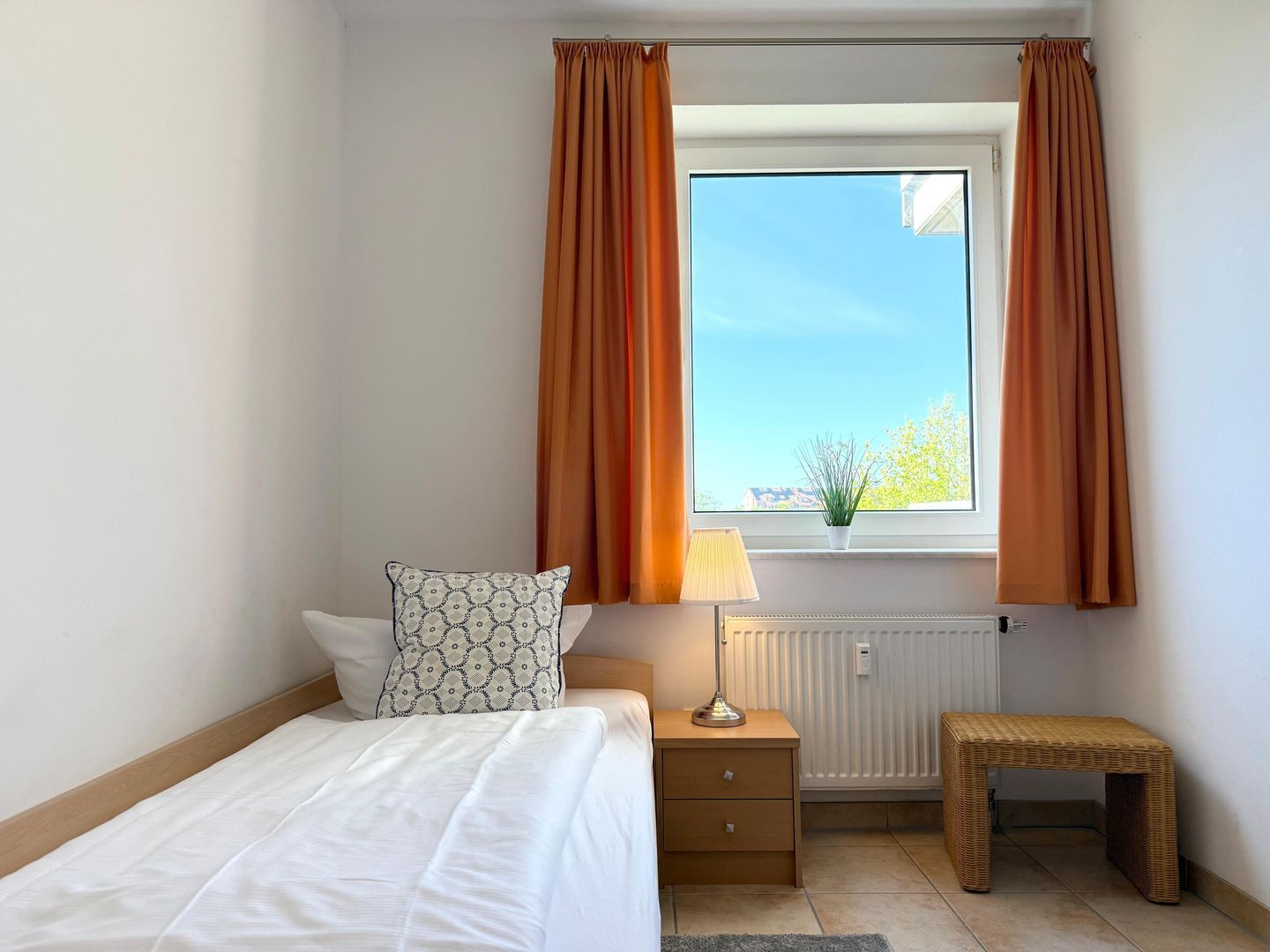 Un dormitorio con una cama, una mesita de noche con lámpara, un pequeño banco, una ventana con cortinas y un radiador.