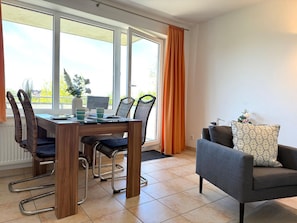 Ein modernes Wohnzimmer mit Essbereich, ausgestattet mit einem Holztisch, Korbstühlen, einem grauen Sofa und einem großen Fenster mit Vorhängen.