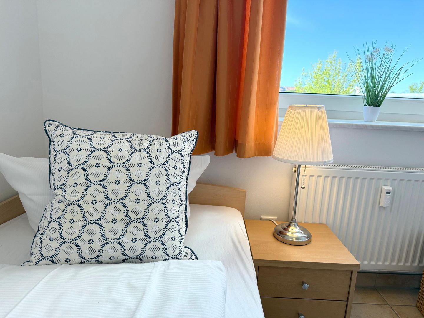 Ein ordentlich bezogenes Bett mit einem gemusterten Kissen, ein Nachttisch mit Lampe und Blick ins Freie durch ein mit Vorhängen drapiertes Fenster.