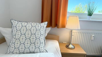 Ein ordentlich bezogenes Bett mit einem gemusterten Kissen, ein Nachttisch mit Lampe und Blick ins Freie durch ein mit Vorhängen drapiertes Fenster.