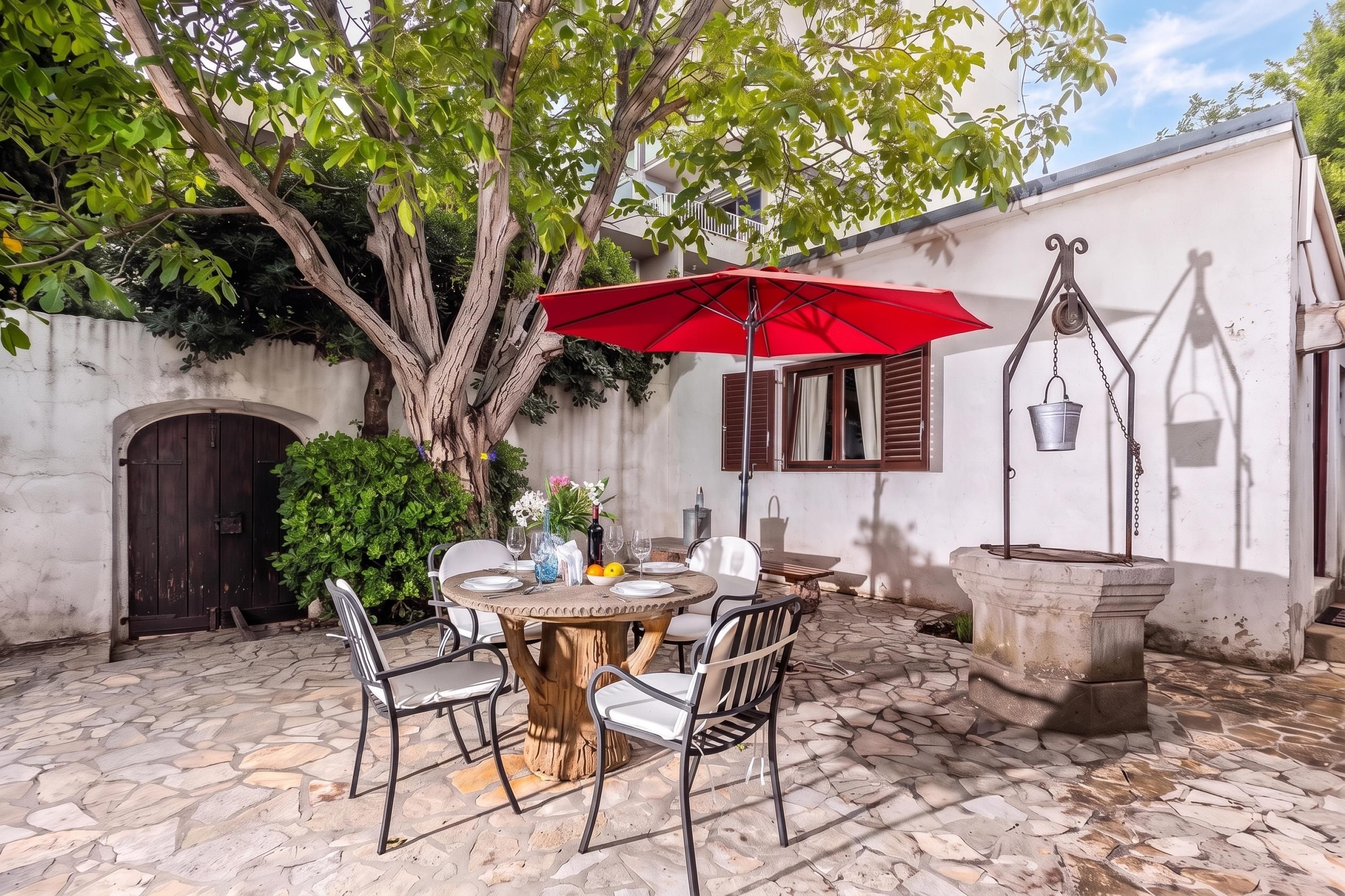 An outdoor patio with a round table, chairs, a red umbrella, and a stone fountain.