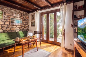 A cozy living room with a stone wall, a green sofa, a wooden table with breakfast items, and a television displaying a mountain landscape. - Lovely Holiday Home By The Sea, Selce, Croatia (Selce)