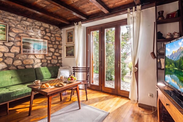 A cozy living room with a stone wall, a green sofa, a wooden table with breakfast items, and a television displaying a mountain landscape. - Lovely Holiday Home By The Sea, Selce, Croatia (Selce)