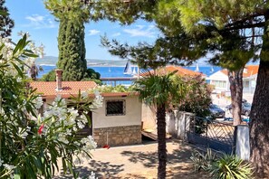 Une propriété située sur la côte, dotée d’un mur en pierre, d’un petit jardin agrémenté de fleurs blanches et offrant une vue sur la mer, où l’on peut apercevoir des voiliers.