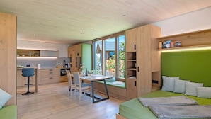 Une chambre moderne avec un lit vert, des meubles en bois, un coin repas et une vue dégagée grâce à de grandes fenêtres.