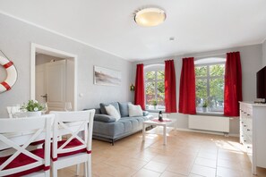 Un salon comprenant un canapé, des fauteuils, une table, une fenêtre avec des rideaux, un radiateur et un luminaire au plafond.