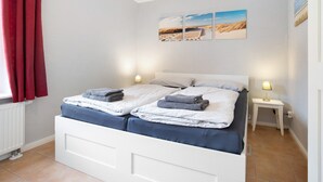 Una cama doble con dos mesitas de noche, una ventana con cortinas rojas y un cuadro que representa una playa.