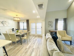A modern living room with a sofa, dining table, and chairs. There is a large window with blinds, a ceiling vent, and a wall-mounted artwork.