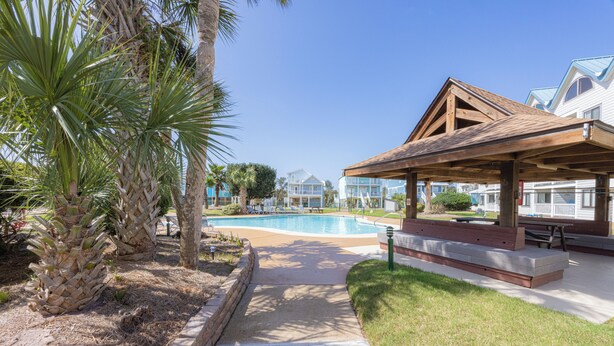Un área de piscina con muelle de madera, palmeras y una zona de estar cubierta.