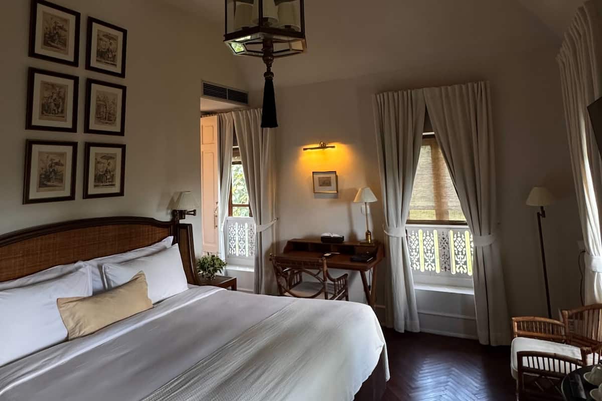 Habitación de hotel con una cama grande, un escritorio y una silla. En la pared hay cuadros enmarcados, una lámpara de techo y cortinas en las ventanas.