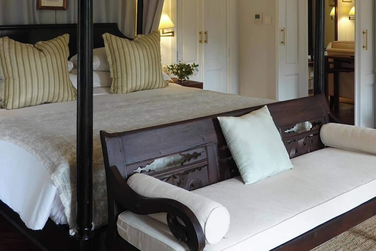 Una cama con dosel y cuatro columnas, con estructura de madera, un sofá con cojín y una mesita de noche con lámpara.