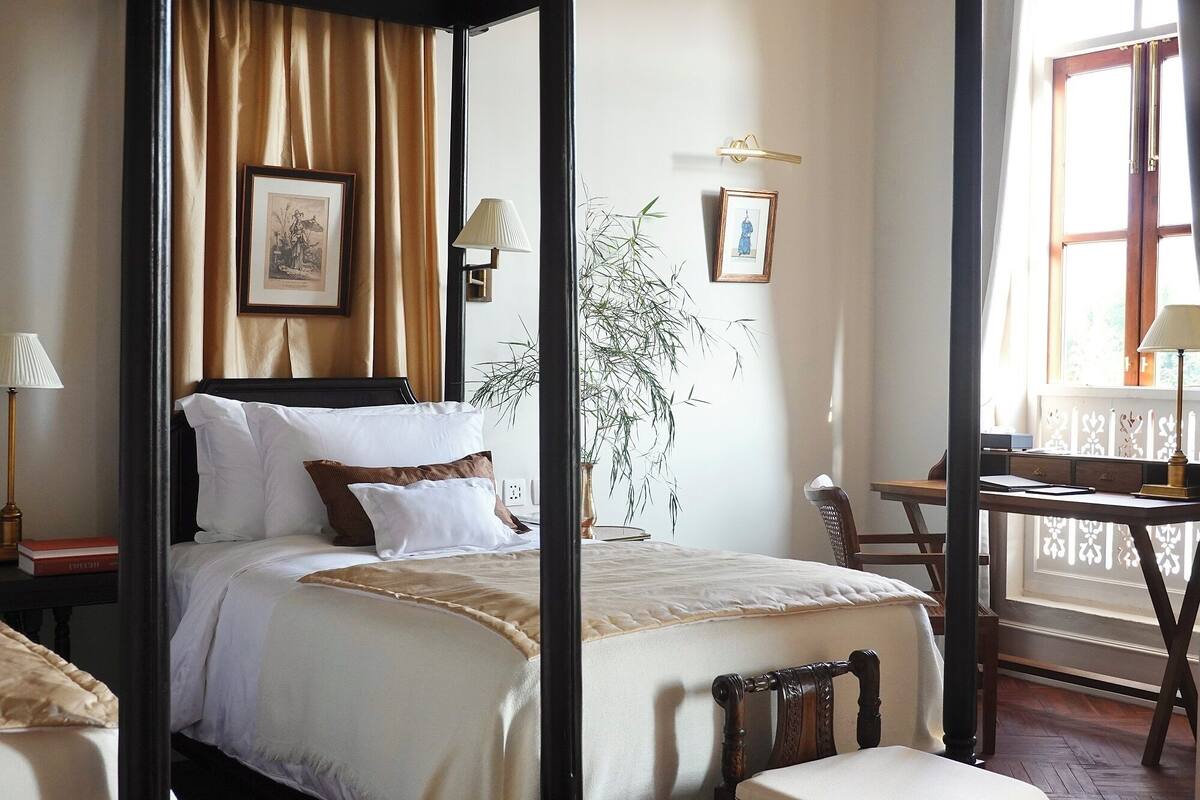 Habitación de hotel con cama con dosel, un escritorio y una silla. Hay dos lámparas a cada lado de la cama.